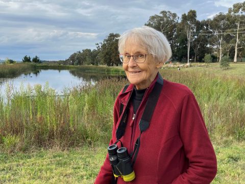 A smiling women standing next to a wildlife pond area (Mawson Ponds) and grasslands, wearing bird watching binoculars